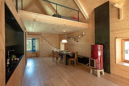 Loft mit heller Holzverkleidung, Esstbereich und roter Kaminofen als Farbakzent, darüber Galerie-Schlafbereich