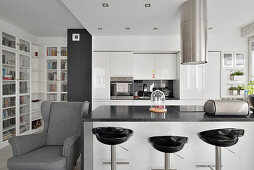 Modern eingerichtete Küche mit Barhockern, grauem Ohrensessel und Bücherregal