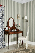 Dressing table with oval mirror and antique chair in front of striped wallpaper