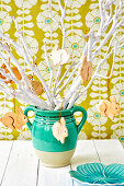 White branches with wooden bunny pendants in a green vase in front of patterned wallpaper