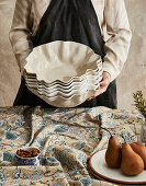 Person in apron with stacked ceramic bowls behind patterned tablecloth