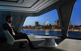 Mann sitz in einer modernen virtuellen Wohnung und schaut auf die Hafenskyline, Hamburg, Deutschland