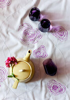 Still life with yellow teapot and purple glasses on tablecloth