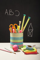 Homemade pen holder made from a tin can on a desk in front of a blackboard with ABC lettering