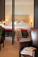 Brauner Lederesessel vor offener Tür und Blick ins Schlafzimmer auf Bett mit hohem Kopfteil und Nachttisch Beleuchtung