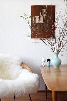 Chair with white animal skin next to a 50's style wooden table and flower vase in front of a wall with a poster hanging on it