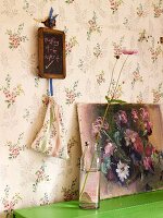 Vase mit Blume, Bild mit Blumenstilleben auf grüner Ablage und kleine Kreidetafel an einer tapezierten Wand mit Blumenmuster