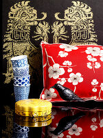 Kissen mit floralem Bezug, Vogelfigur, Schatulle, Pappbecher vor asiatischem Wandbehang