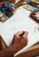 Nahaufnahme einer zeichnenden Männerhand auf Skizzenblock, Malutensilien unscharf im Hintergrund