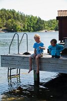 Two children sitting on wooden jetty on shore of lake playing with wooden toy boat
