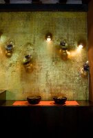 Bowls on black sideboard against artistically decorated wall with integrated water spouts, vases and sconce lamps