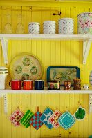 Storage jars and colourful, vintage, enamel mugs on shelves on yellow-painted wall