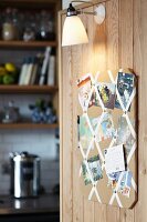 Wandboard mit Postkarten und Fotos, beleuchtet von einer Lampe, auf Holzwand, dahinter Bücherregal und Kaffeemaschine
