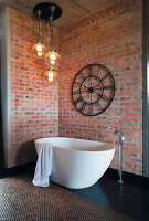 Freestanding bathtub in front of brick wall with large wall clock and globe lights