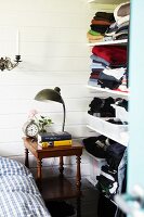 Lamp on antique side table next to open-fronted shelves of clothes on white wooden wall