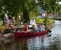 Rotes Kanu auf Fluss mit zwei Frauen an Bord und Kindern an sommerlichem Badesteg