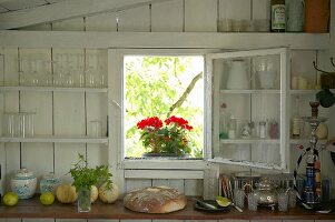 Vintage Holzwand mit offenem Fenster und Blick auf rote Geranien, Regalbretter mit Gläsern und Küchenarbeitsplatte mit Brotlaib