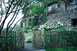 Knüppelzaun mit offenem Tor, weiße Fliederbüsche vor altem Landhaus teilweise mit Naturstein und Schindel Fassade