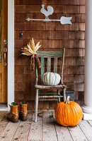 Kürbisse, getrocknete Maiskolben, rustikaler Holzstuhl und Stiefeln auf herbstlicher Veranda