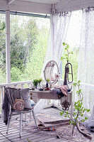 Oval mirror and trumpet used as vase on dressing table and vintage chair on roofed terrace with curtains
