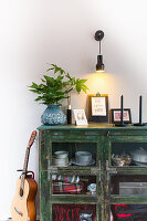 Grüner, halbhoher Vitrinenschrank, Vase mit Blätterzweig, Bilder und Kerzen, darüber Wandleuchte