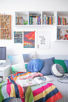 Colourful knitted blanket and cushions on bed below bookshelves
