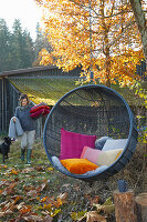 Colourful cushions in hanging chair in autumnal garden: woman in background carrying cushions and blanket