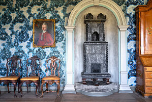 Antiker Gusseisenofen, Portraitmalerei und Stühle vor Wand mit blau gemusterter Tapete