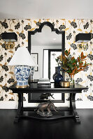 Schwarzer Konsolentisch mit Tischleuchte und Blumenstrauß vor Tapete mit floralem Design und Wandspiegel