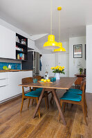 White fitted kitchen, dining table and chairs with blue seat cushions below yellow pendant lights