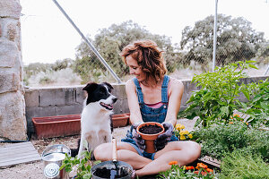 Smiling woman holding flower pot looking at border collie while sitting vegetable garden