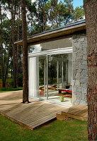 Modernes Haus aus Glas, Holz und Stein in einer Waldlandschaft; Blick von einer Veranda mit Sonnenliegen ins Schlafzimmer