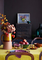 Esszimmer mit dunkelvioletten Wänden und einem herbstlich gedeckten Tisch mit goldgelber Tischdecke und Blumen und Obst der Saison