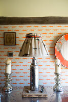 Upcycled table lamp made from old thermos flask flanked by candlesticks on table in front of wallpapered wall