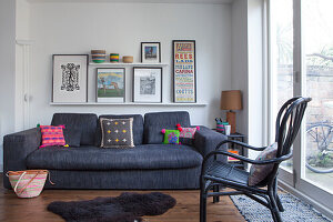 Blue sofa with scatter cushions below collection of pictures on shelves; black rattan chair in the foreground