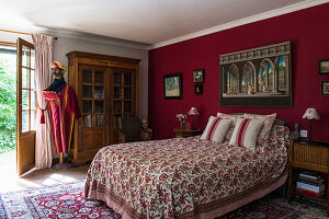 Double bed with floral bedspread, antique wardrobe and mannequin in Afghan costume in bedroom with red walls