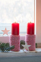 Two burning red candles on spools of yarn, fir branch and stars on windowsill