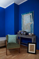 Upholstered chair with cushions in front of console table and painting in room with blue walls
