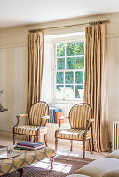 Pair of striped antique style chairs in window of sitting room with Regency style ottoman and linen floor length curtains