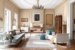 Upholstered sofas, chaise lounge, and antique wardrobe in a living room with opulent chandelier and beige walls