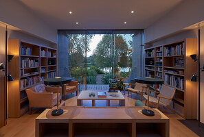 Beleuchtetes Wohnzimmer mit Bücherregalen und Sitzmöbeln, Blick durch Terrassentür aus Glas auf Fluss