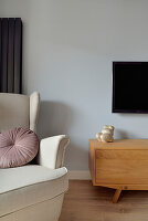Light-coloured wing chair with cushion next to lowboard and TV on the wall