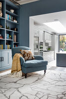 Blaue Regalwand, davor farblich passender Sessel im Wohnzimmer, im Hintergrund helle Küche