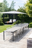 Large wooden terrace with dining table, white chairs, umbrella and garden pavilion in the background