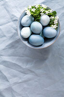Blue Easter eggs and white flowers in a ceramic bowl on a blue linen tablecloth