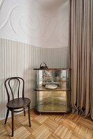 Glass cabinet with biscuits and wooden chair next to pleated curtain in three-room apartment