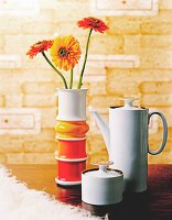 Coffee pot, sugar bowl and vase with gerbera daisies in orange and yellow in front of wallpaper