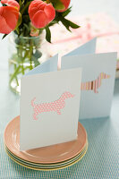 Homemade greeting cards with dog motif on stack of plates and table, flowers in the background