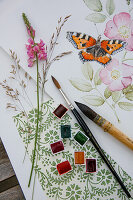 Watercolour painting of butterfly on dog rose with painting utensils